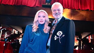 Madonna and Graham Norton look to the camera. They stand side-by-side in front of a stage and technical equipment. 