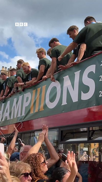 An open top bus with rugby players drivers past cheering crowds