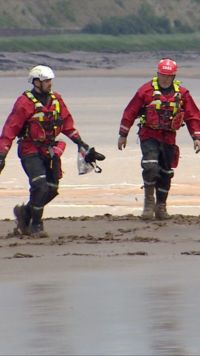Two people walking in safety uniform on sand with water behind them.