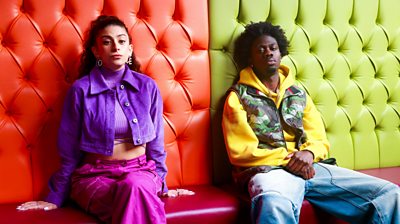 Two teenagers sit on a sofa which is half red-half green. They are in colourful outfits and have serious expressions.