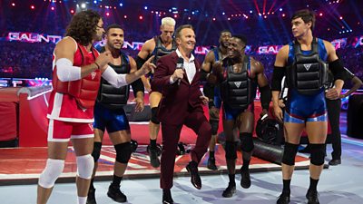 Presenter Bradley Walsh marches forward in a burgundy suit, alongside a line up of Gladiators contestants. The contestants are all wearing shorts and vests underneath body protection gear.