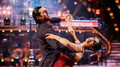 Balvinder Sopal and Julian Caillon dance the Waltz. Julian is wearing a dark pinstripe suit with black slick back hair, and Balvinder wears a red long, sparkly ballgown.