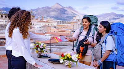 Two women with backpacks standing beside a table outdoors, speaking with another woman, who is handing them a map against a mountainous backdrop.