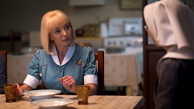 A woman in nurse's uniform seated at a table in a kitchen, speaking to someone off camera.