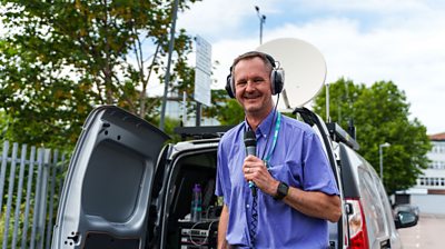 Jeremy Buxton holding a microphone while standing beside broadcast equipment outdoors.