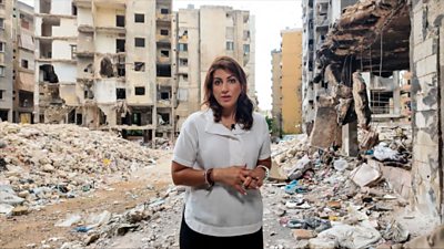 Carine Torbey, with brown hair and wearing a white short sleeve shirt, reports from Lebanon amidst a background of destroyed high rise buildings and debris.