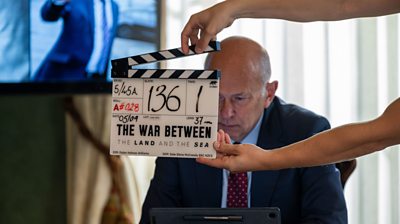 A clapperboard is held in the foreground with production details for The War Between the Land and Sea. Prime Minister Harry Shaw (Vincent Franklin) sits in the background looking at an iPad, wearing a suit.