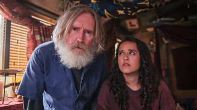 Michael Sleep (Pearce Quigley) has long grey hair and a grey beard and looks at the camera in thought, wearing a blue overall shirt whilst Kacey (Lauren Patel) looks at him. Kacey has burgundy long sleeve shirt and brown curly hair.