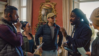 Three people standing in front of a pink wall whilst filming, with one person wearing a cap and glasses speaking to another.