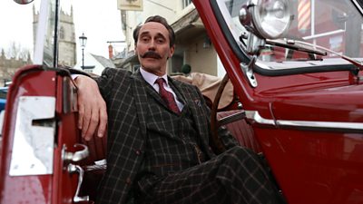 Guy Henry in a red sports car in a three-piece suit with a bug moustache