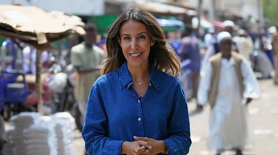 Broadcast jounralist Nawal Al-Maghafi reports from Sudan for Global Eye. She stands in front of a blurred market background, with hands clasped in front and a blue button up shirt. She has long brown hair.