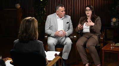 Victoria Derbyshire with her back to the camera, interviews Amy and John Hunt opposite her. Amy wears a brown suit and John wears a grey suit.