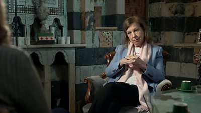 Chief International Correspondent Lyse Doucet during an interview in Syria. Lyse sits in a living room, on a brown armchair. She has her hands clasped in front of her with a pale blue blazer and pink scarf on. She has brown hair.