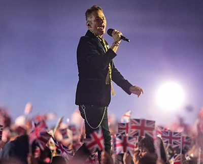 Joseph Washbourn walks along a stage singing surrounded by fans waving union jacks. He holds a microphone and is wearing a dark suit.