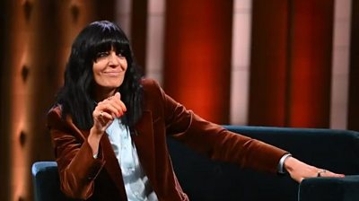 Claudia Winkleman looks sideways on a sofa chair in a TV studio. She is smiling and wears a copper velvet blazer and pale blue shirt.