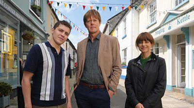 Actors Kris Marshall and Dylan Llewellyn stand facing the camera on a Cornish street with bunting.