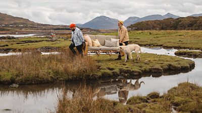 Banjo and Ro walk over across a moor on a Scottish island carrying a sofa. A white dog walks aside them.