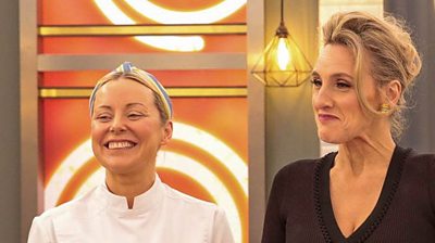 Anna Haugh and Grace Dent stand laughing in the Masterchef studio in front of lights and Masterchef branding. Anna wears white chef's uniform and Grace wears a black v neck dress.