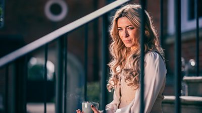 Fran (Eve Myles) sitting on steps, holding a cup and looking into the distance. She has long, blonde curly hair and wears a white blouse and beige waistcoat.