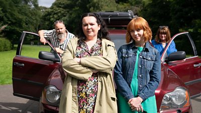 Nina (Ashley Storrie) sits on a red car bonnet with her arms folded, wearing a floral dress and beige coat. Next to her is Evie (Kat Rooney)wearing green jumpsuit and denim jacket. In the background, both car doors are open. Diane (Sally Howitt) and Ade (Greg Hemphill) are in the background.
