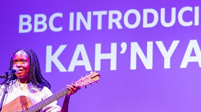 Musician Kah'nya stands in front of a BBC Make a Difference Award screen with a brown guitar and sings into a microphone.