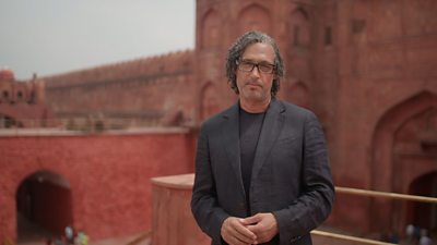 David Olusoga stands outdoors in a historial site looking into the camera. He wears a black blazer and a grey t shirt, and rectangle glasses.