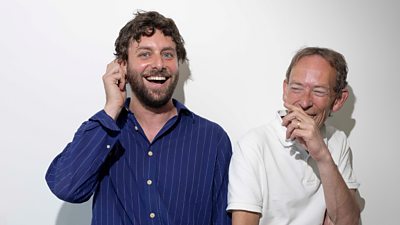 A man in a blue striped shirt and a man in a white polo shirt laugh as they pose for a photo against a white backdrop