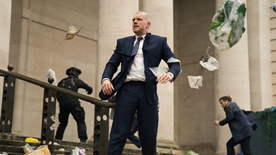Barclay Pierre-Dupont (Russell Tovey) stands on the steps to a large building in a navy suit as papers swirl around him