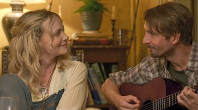 Nell Mortimer (Carey Mulliagn) smiles in a pair of dunagrees looking at Herb McGwyer (Tom Basden) who plays the guitar to her. He wears a checked flannel shirt.