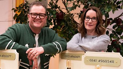 Alan Carr and Susie Dent stand in amidst plinths with different coloured telephones and lightbulbs on. There is a fruit tree in the background.