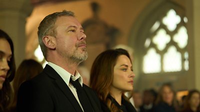 A man in a suit sits looking forward at a funeral. He's flanked by two women in the congregation.