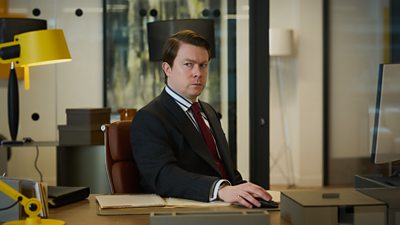 A man sits behind a desk in an office, looking suspiciously at something off screen