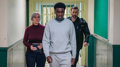 Christopher (Chukwubuikem Molokwu) wears a grey tracksuit and walks down a corridor, followed by Marie-Louise O’Dell (Siobhan Finneran) and Bobby Bailey (David Tennant). 