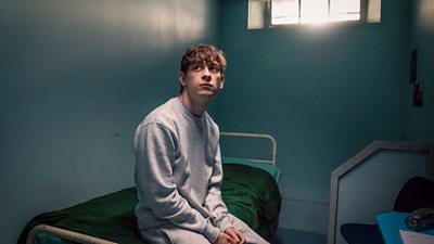 James (Louis McCartney) wears a grey tracksuit and sits on the edge of a bed in a prison cell. 