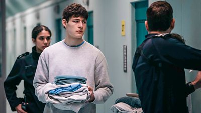 Ollie McNulty as Peter. Wearing a grey tracksuit, he carries a pile of clothes towards a prison cell. Prison officers stand nearby. 