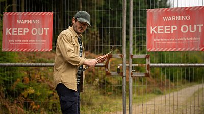 Iwan Rheon wearing a shirt and a cap stood next to a road blocked by a gate. On the gate are red warning signs saying Keep Out