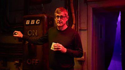 Louis Theroux looks earnestly to camera as he stands with one hand on a giant lever, which represents an on/off switch for the BBC