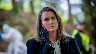A woman in a suit wearing black gloves holds a phone to her ear and looks at the camera. In the background is a blurred crime scene in a forest