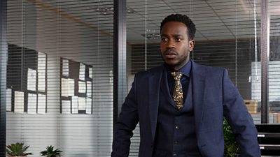 A man wearing a blue suit and tie standing in an office looking intently away from camera 