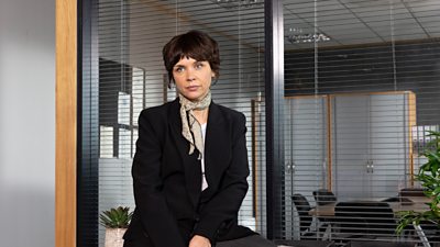 A woman wearing a suit perched in a generic office room looking away from the camera 