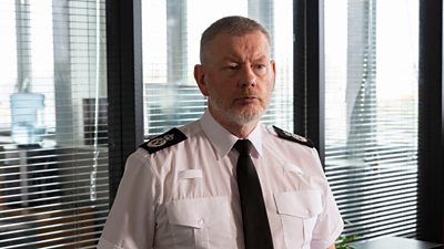 A man in a police uniform looking stern in a generic office room