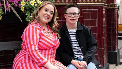 A woman and a man - Charley Marlowe and George Webster - sit on a bench outside the Queen Vic pub, smiling