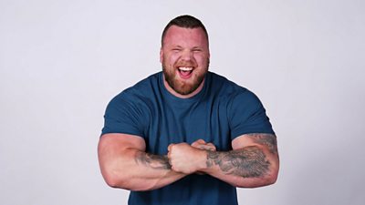 A man wearing a blue t shirt flexing his muscles and laughing at the camera