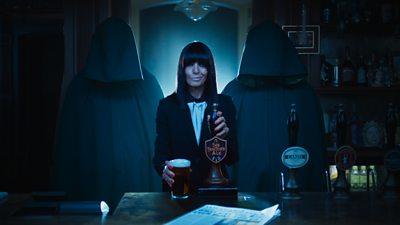Claudia Winkleman stands behind the bar in the Queen Victoria pub serving Traitors Ale, while flanked by two hooded figures