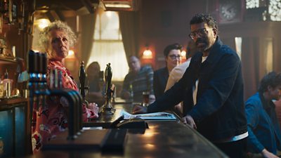 Romesh stands at the bar in the Queen Victoria pub, where longstanding barmaid Tracey is serving. They both look to camera as Ian Beale and Sue Perkins watch on.