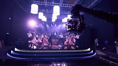 A camera on a crane pans across a scene where Romesh stands on a raised podium, surrounded by Strictly Come Dancing dancers