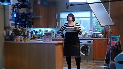 Ruth Jones stands in a kitchen with a cup in hand, filming scenes as Nessa from Gavin & Stacey