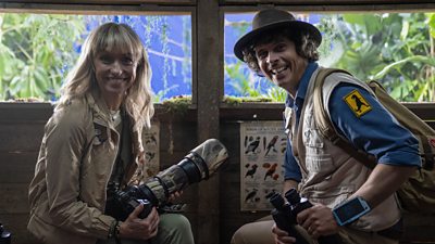 Nature presenters Michaela Strachan and Andy Day sit in a bird hide, smiling to camera