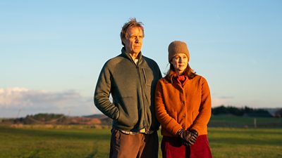 Scene from Onwards and Sideways. Rhys Ifans and Laura Linney stand outdoors in a field. They both wear warm clothing and are illuminated by warm sunlight. They look at something offscreen.