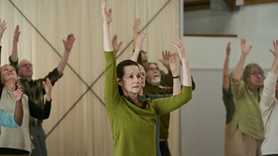 Scene from Onwards and Sideways. Emma Dretzin (Laura Linney) performs a yoga pose in a studio full of other yoga class members. She looks straight ahead with a worried expression.